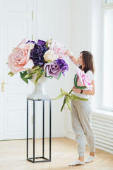 Indoor shot of female florist holds rose, creats beautiful bouquet, decorates room, owns flower shop, dressed in casual clothes, makes composition. Occupation and floristy concept. Designing.