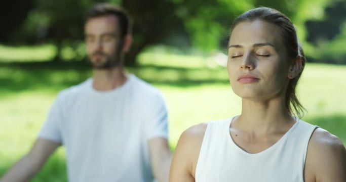 Close up of the pretty young woman meditating while doing her morning yoga in the park. Attractive man on the background.