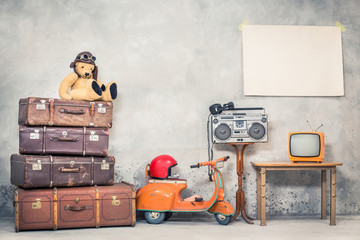 Retro Teddy Bear toy in aviator's hat, aged travel valises, children's pedal scooter, helmet, cassette boombox with headphones, TV, paper poster blank on concrete wall. Vintage style filtered photo