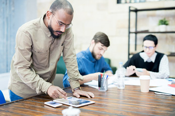 Fototapeta premium Serious pensive hipster manager in glasses synchronizing tablet and smartphone while transferring files, he standing at table in board room