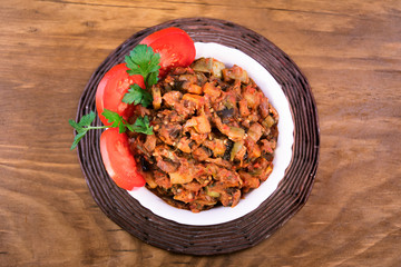 Stew of roasted vegetables in a white bowl with tomatoes and parsley, top view, wooden background.