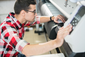 Serious thoughtful handsome young engineer in shirt examining wide format printer while holding...
