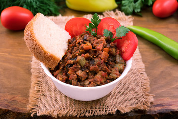 A portion of vegetable stew with eggplant, a piece of white bread with tomato slices and parsley in a white bowl.
