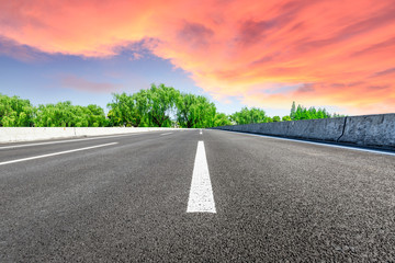 Empty asphalt road and green forest with colorful clouds at sunset
