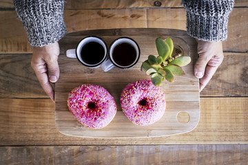 above view of pair of caucasian female hands taking a wood plate with two lack italian coffee and a couple of pink sugar donuts. wood table in background. morning and no diet concept happy lifestyle