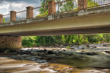 Smooth flowing river going under a stone and steel bridge with the sun reflecting off the water and rocks and moody clouds in the sky - obrazy, fototapety, plakaty