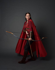 full length portrait of brunette girl wearing red medieval costume and cloak, holding a bow and...