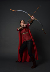 full length portrait of brunette girl wearing red medieval costume and cloak, holding a bow and...