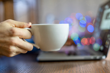 male hand holding a white cup with hot drink on work table whily typing on laptop keyboard f