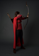 full length portrait of brunette girl wearing red medieval costume and cloak, holding a bow and...