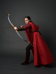 full length portrait of brunette girl wearing red medieval costume and cloak, holding a bow and...