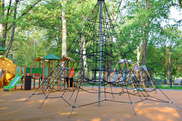 Fototapeta premium Modern playground with climbing net swings and slides with soft protective rubber coating in city park with old trees and pavement pathway in summer day. Children activities outdoors concept.