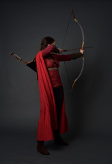 full length portrait of brunette girl wearing red medieval costume and cloak, holding a bow and...