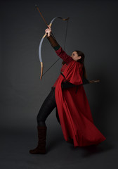 full length portrait of brunette girl wearing red medieval costume and cloak, holding a bow and arrow. standing pose on grey studio background.