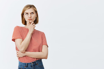 Thinking, making important and hard choice in mind. Thoughtful intense attractive and young girl with fair hair, touching chin, frowning while staring up at ceiling, hesitating over gray background