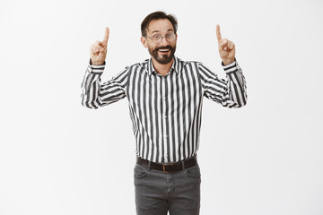 Naklejka premium Look upstairs dear guest. Charming and hospitable man with beard in glasses and striped shirt, pointing up with raised fingers and smiling broadly while advertising or promoting his campaign