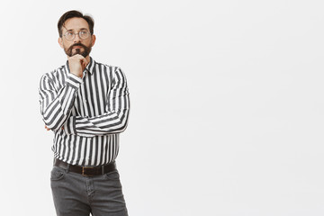 Charming and creative strategist thinking of new invention, standing in mindful and thoughtful pose, leaning on fist and gazing up with slight smile, posing dreamy over gray background