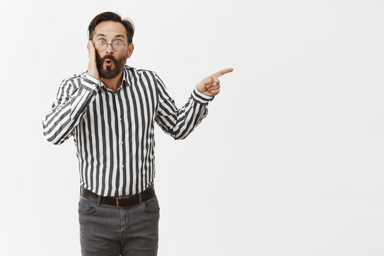 Portrait Of Impressed And Thrilled Gay Man With Beard And Moustache, Folding Lips In Curious Emotion And Pointing Right, Holding Palm On Cheek, Being Surprised And Shocked Person Showed Up On Party