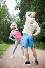 Active mature couple in sportswear doing physical exercise in park among green trees