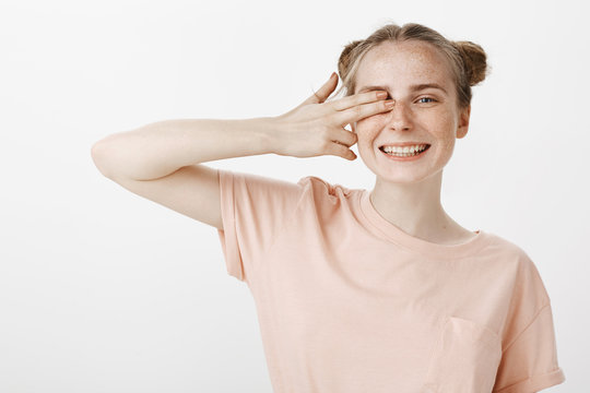 I am watching you. Portrait of carefree emotive young woman with freckles and blue eyes, smiling broadly while covering one eye with two fingers, being in gread friendly mood over gray background