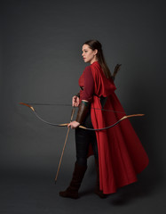 full length portrait of brunette girl wearing red medieval costume and cloak, holding a bow and...