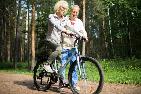 Aged blonde woman riding bicycle in the forest while her husband teaching her