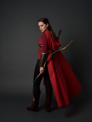 full length portrait of brunette girl wearing red medieval costume and cloak, holding a bow and...