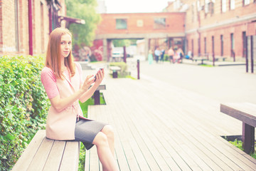 Beautiful young girl sitting on a wooden bench on an open Looking cell phone smiling Sunny day green bushes lifestyle Selective focus toning