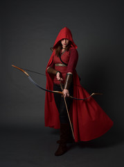 full length portrait of brunette girl wearing red medieval costume and cloak, holding a bow and...