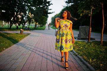 Fototapeta premium Cute small height african american girl with dreadlocks, wear at coloured yellow dress, posed at sunset.