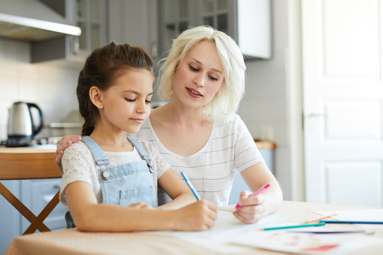 Young blonde female looking at her daughter drawing while both sitting in the kitchen
