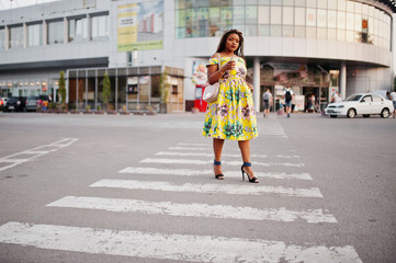 Cute small height african american girl with dreadlocks, wear at coloured yellow dress, walking at crosswalk against trade center with cup of coffee at hand.