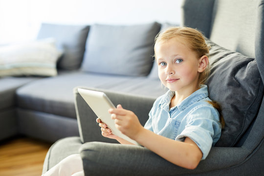 Youthful girl with tablet sitting on sofa in living-room and looking at you while watching online video