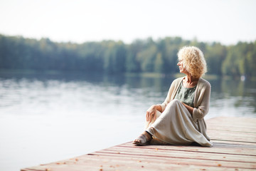 Serious pensive curly-haired mature lady in long skirt sitting on pier and contemplating tranquil...