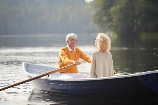 Cheerful excited affectionate senior man in yellow sweater falling in love with beautiful curly-haired lady rowing boat and smiling at girlfriend while enjoying date on boat.
