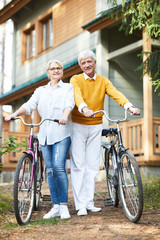 Obraz premium Smiling beautiful senior couple in casual clothing leading active lifestyle: they holding bicycle handles and looking at camera while resting in countryside