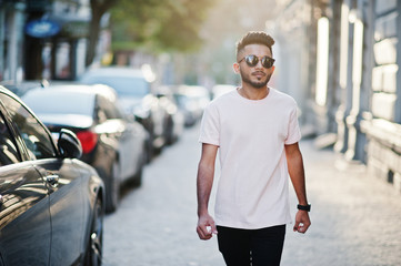 Obraz premium Stylish indian beard man at sunglasses and pink t-shirt. India model walking outdoor sunset at streets of city.