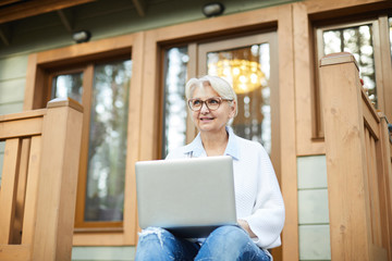 Smiling modern working senior lady in glasses sitting on porch of cottage and looking into distance...