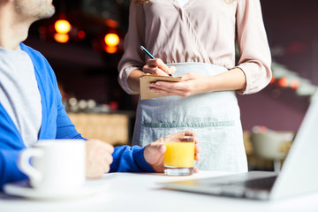 Close-up of unrecognizable waitress in apron making notes while taking order from customer in restaurant