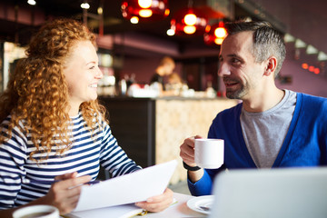 Cheerful successful entrepreneurs sitting at table and discussing papers while planning startup project in cafe