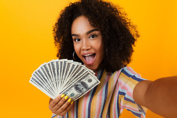 Excited young african woman posing isolated over yellow background holding money make selfie by...