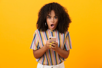 Shocked emotional african woman posing isolated over yellow background using mobile phone.