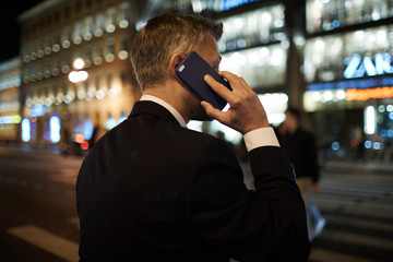 Back of busy employee talking by smartphone while standing by road in the evening