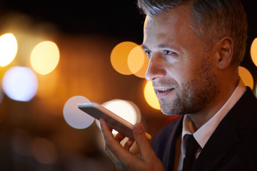 Contemporary businessman recording voice message on smartphone while communicating through mobile...