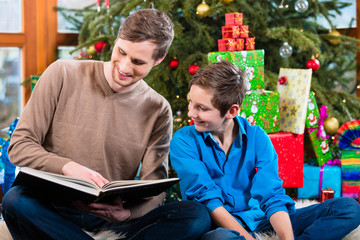 Daddy reading out from book for kid under X-mas tree on Christmas