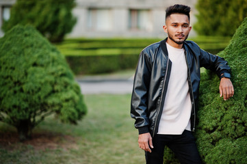 Stylish indian beard man at black leather jacket. India model posed at green garden of city.