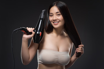 portrait of smiling asian woman with hair dryer looking at camera isolated on black