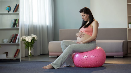 Young pregnant lady listening to her babys beating, sitting on fitness ball