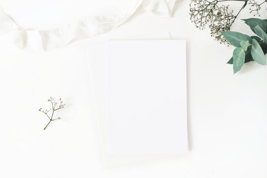 Winter wedding desktop stationery mockup. Blank greeting card, baby's breath Gypsophila flowers, eucalyptus branch and silk ribbon. White table background, top view. Christmas flatlay.