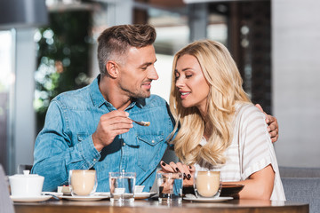 handsome boyfriend feeding attractive girlfriend with sweet dessert at table in cafe